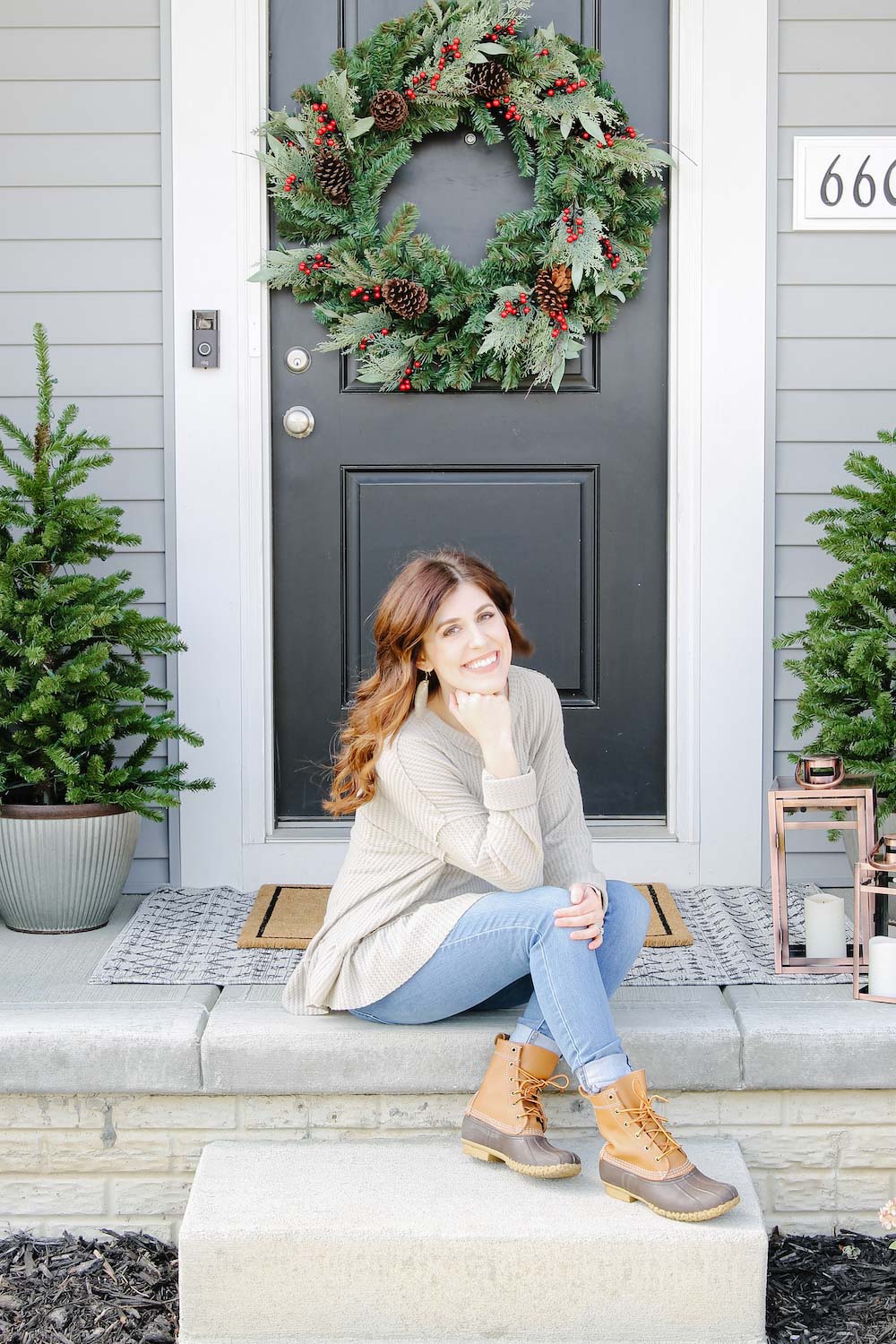 A woman poses on porch in front of oversized holiday wreath and porch trees styled with bronze lanterns