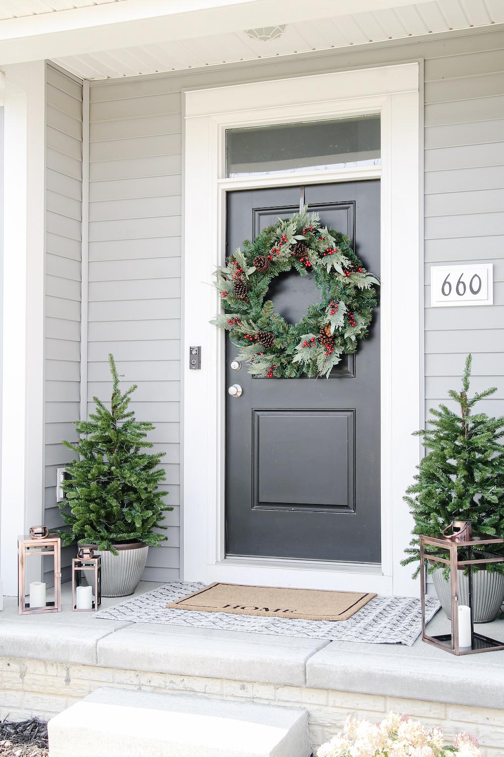 A front porch styled for the holidays with door wreath, porch trees and lanterns shot from right