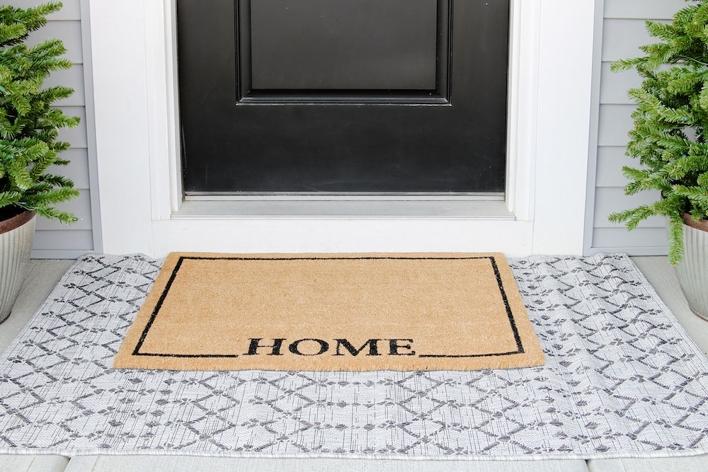 A Home doormat layered on top of a geometric rug between two porch trees and black front door