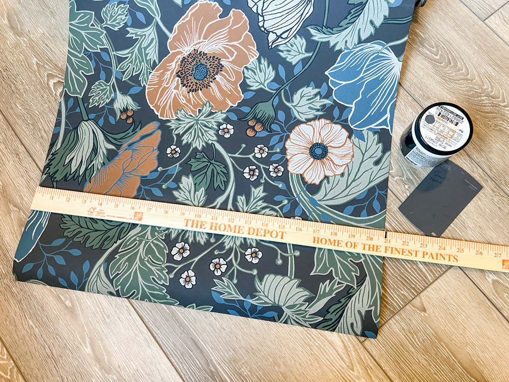 A yardstick on top of a floral wallpaper next to a small container of dark gray paint.