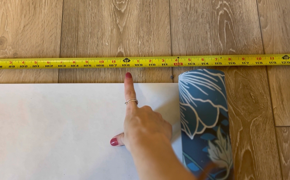 A woman’s hand pointing to a marker on a tape measure next to wallpaper.