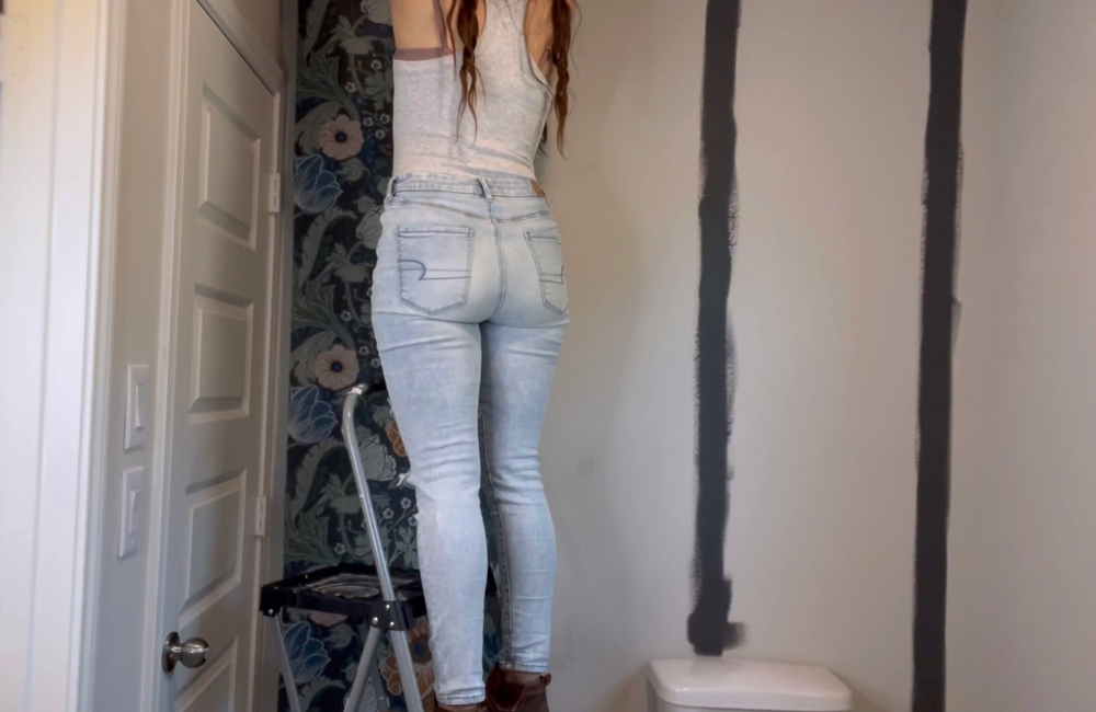 A woman standing on a step stool to apply wallpaper to a wall.