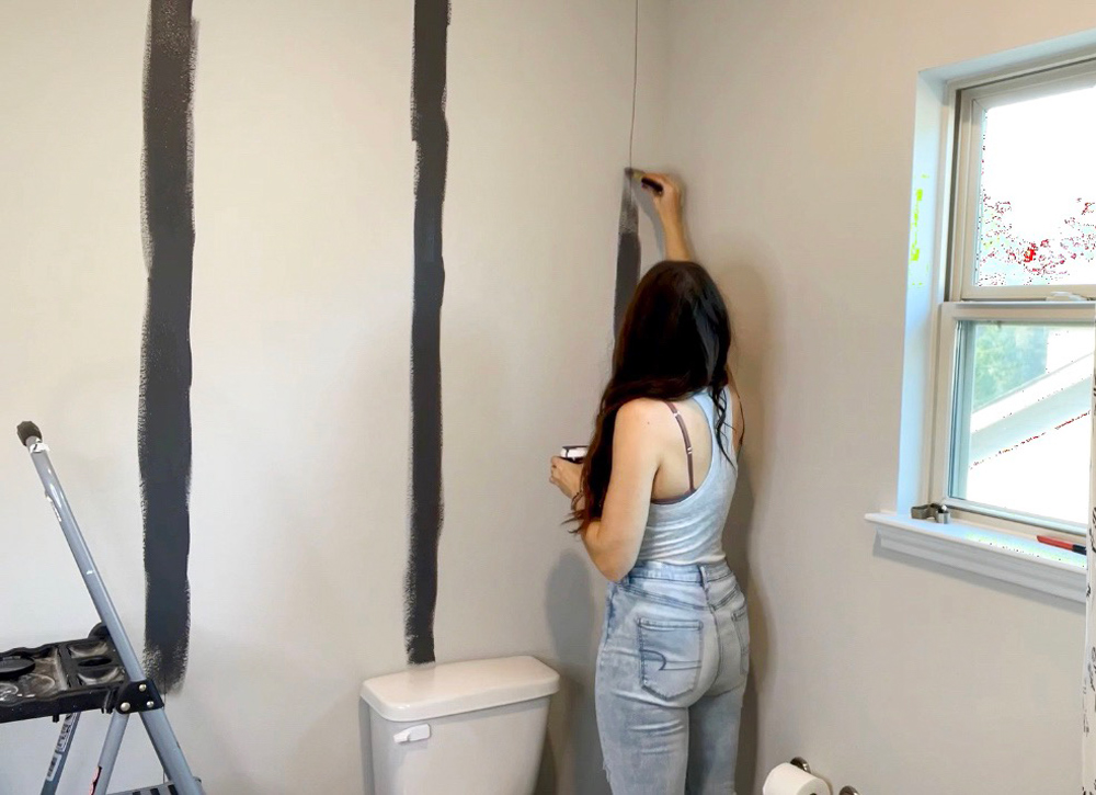 A woman applying dark paint to a wall in vertical stripes