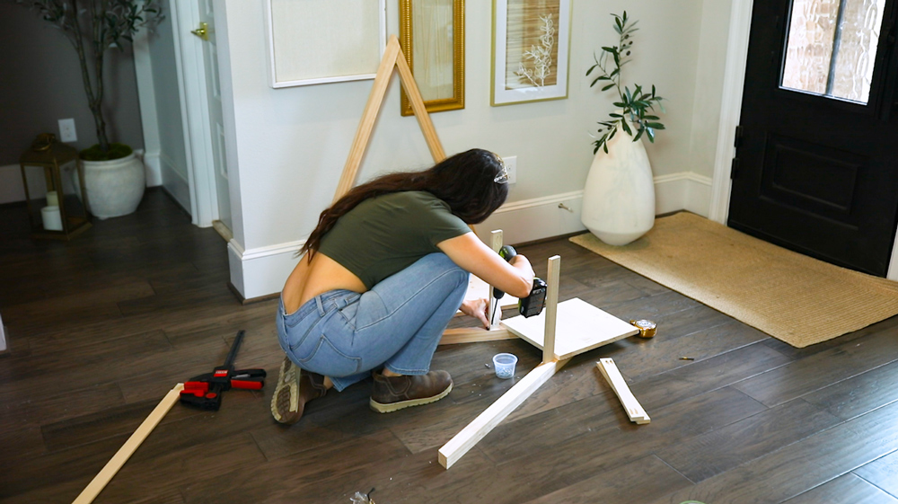 A woman on the ground using a drill to connect side pieces to a wooden frame