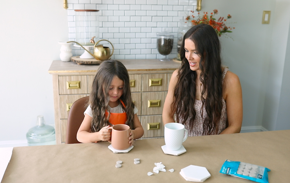 A girl placing a mug on a DIY Tile Coaster while a woman watches.