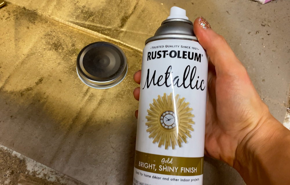 A mason jar lid spray painted gold with a hand holding spray paint canister.