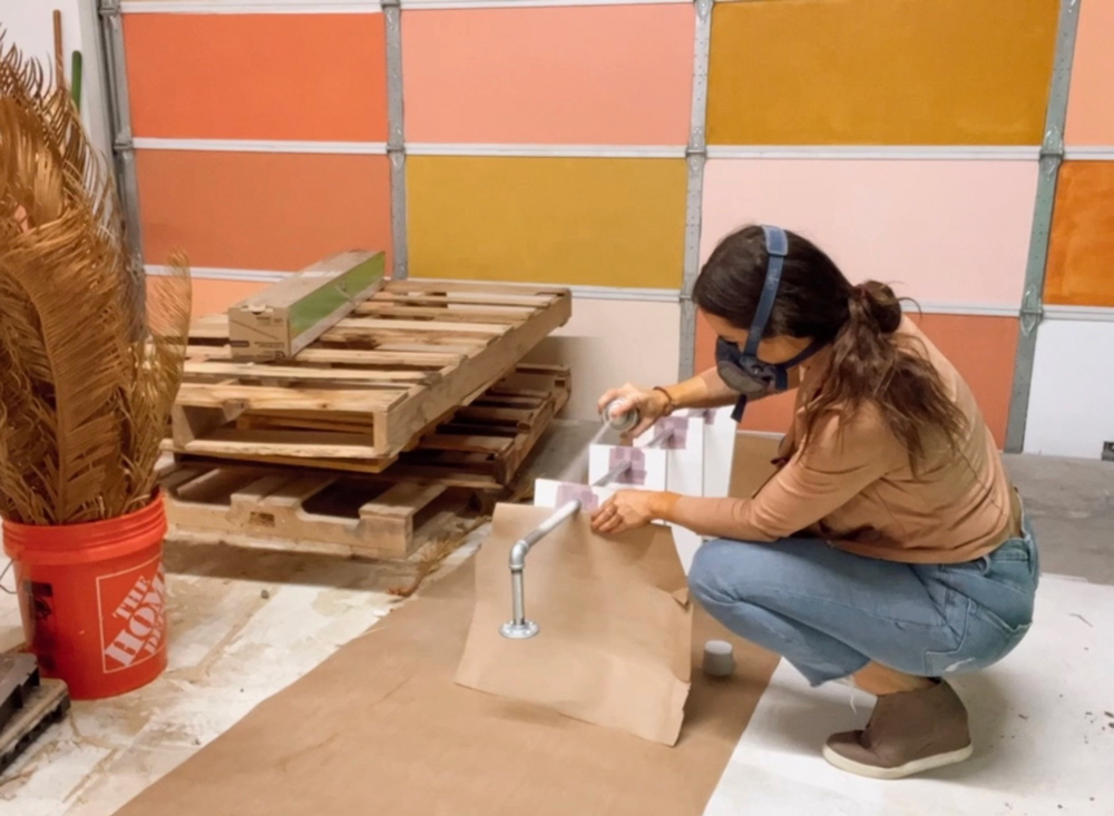 A woman spray painting a DIY plant shelf fixture.