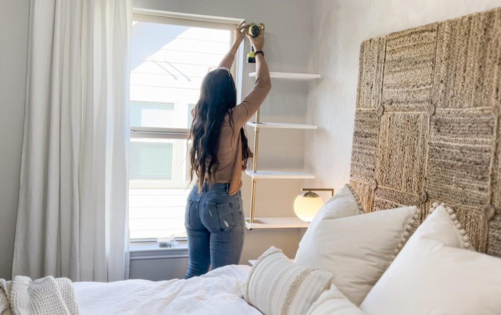 A woman using an electric drill to attach a plant shelf to a wall.