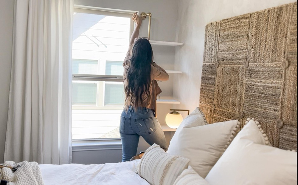 A woman attaching a DIY plant shelf to a wall next to a window.