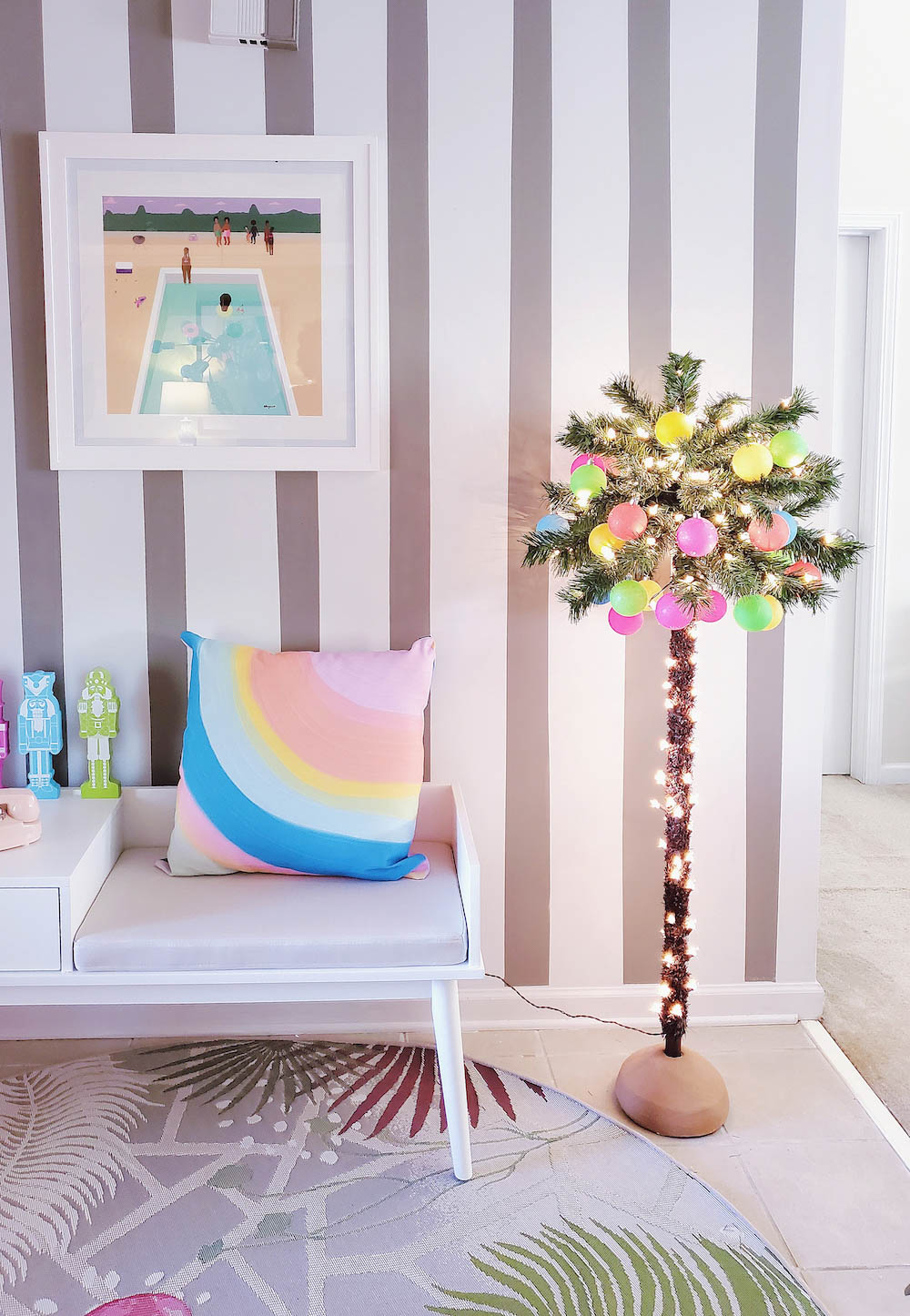 A small indoor palm tree decorated with ornaments and lights positioned next to a bench and in front of a striped wall