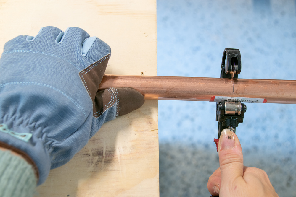 A gloved hand holding a copper pipe while a second hand uses a pipe cutter