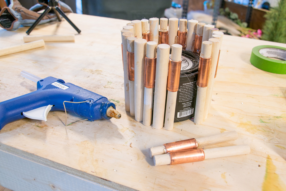  A glue gun lying next to a paint can with wooden and copper dowels attached.
