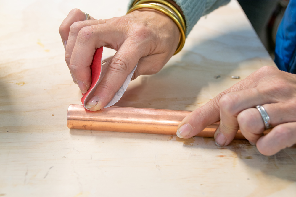 A hand holding a sawed off copper pipe while a second hand uses sandpaper.
