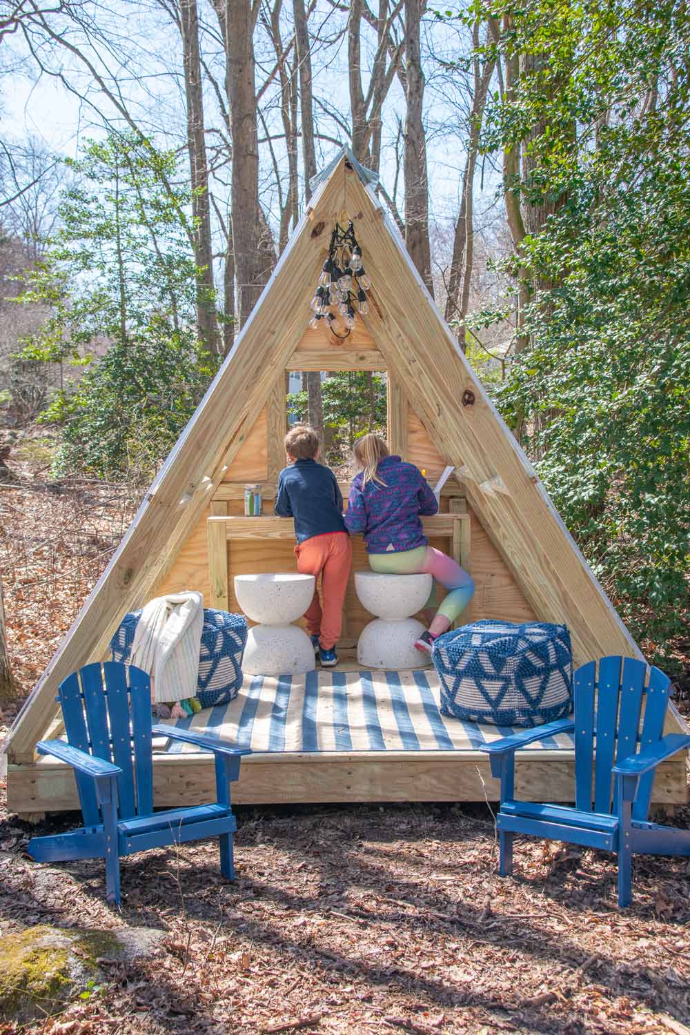 Boy and girl playing in a Luxury A-frame playhouse with two stools, two poofs, and a blue chair.