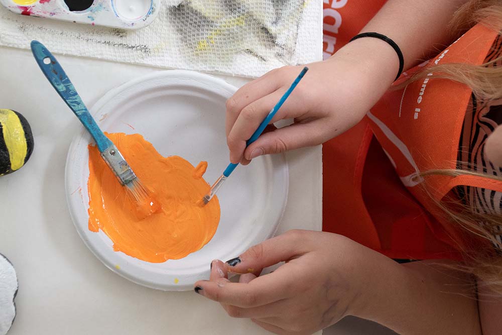A child spreading orange pain on a paper plate with two paint brushes.