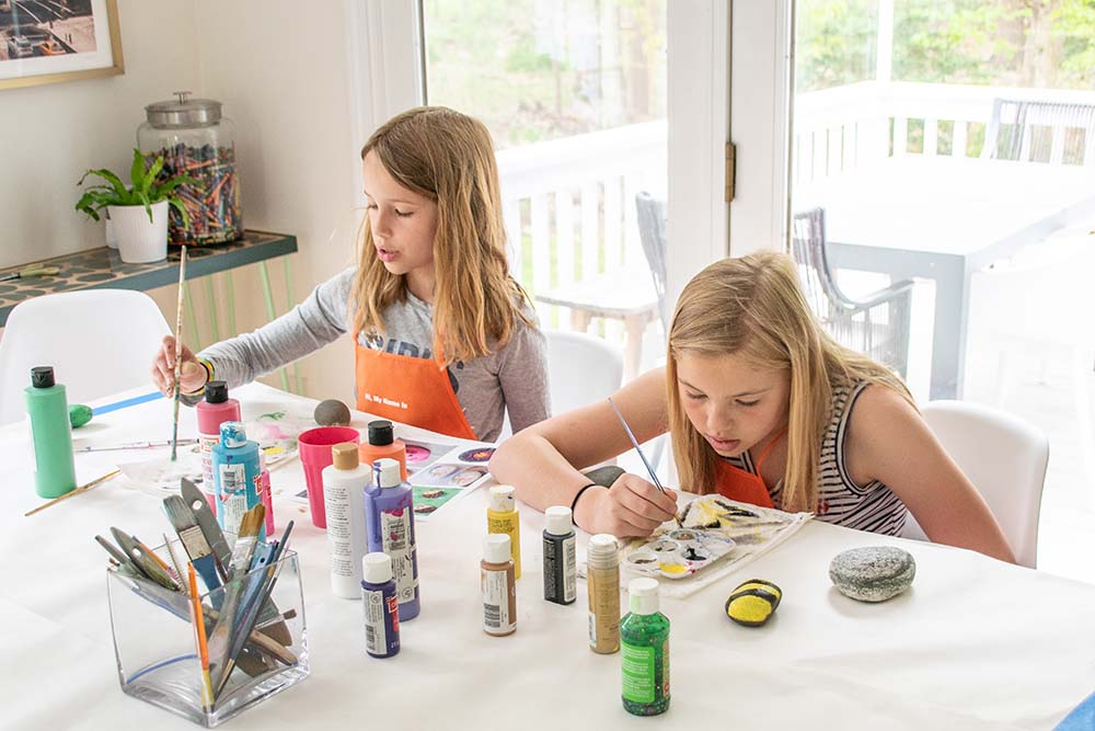 Two children using various paints and brushes to decorate small rocks.