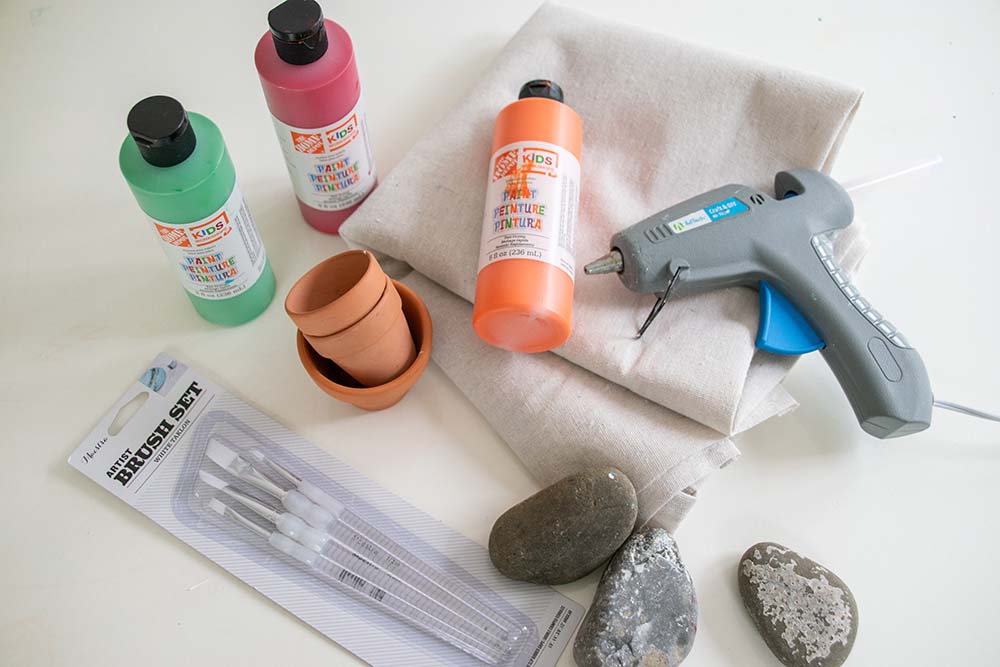 A set of art supplies for the project; a hot glue gun, small rocks, paintbrushes, and paint.