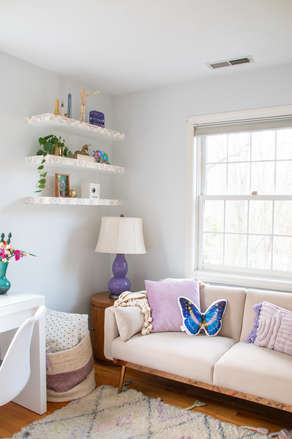 Corner of a kid's bedroom with three decorated floating shelves, basket, table with a purple lamp, and loveseat with throw pillows.