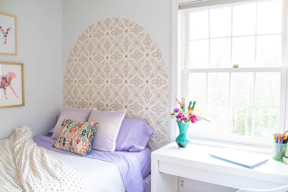 Bedroom with bed with purple sheets, wallpaper headboard, and white desk.