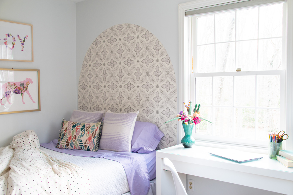 Kids bedroom with gold-framed photos, bed with lavender sheets, and wallpaper headboard.
