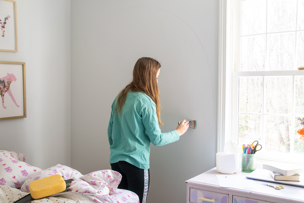Girl using a brush to paint wallpaper paste onto the wall. 