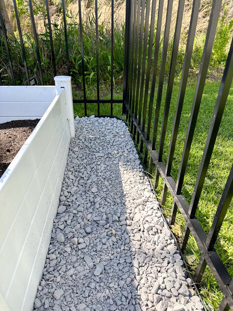 Light grey pebble stones laid within Vigoro No Dig Edging between white raised garden bed and black fence.