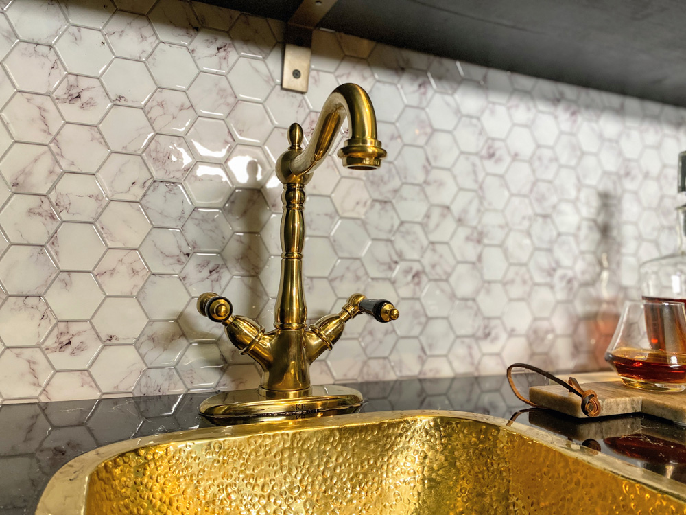 Gold faucet and sink base featured in front of marble mosaic tile backsplash.