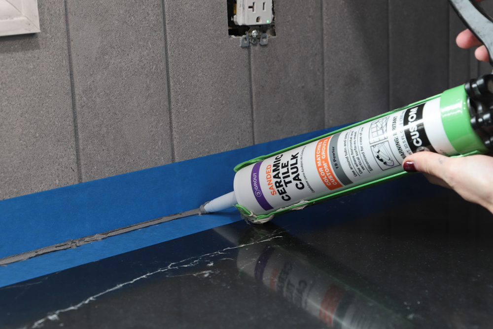 Person using a caulking gun to the taped-off seam of the wall.