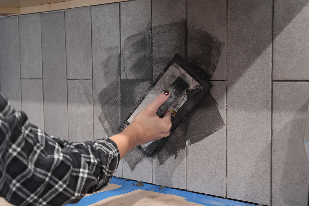 A person applying dark grout to gray tiles.