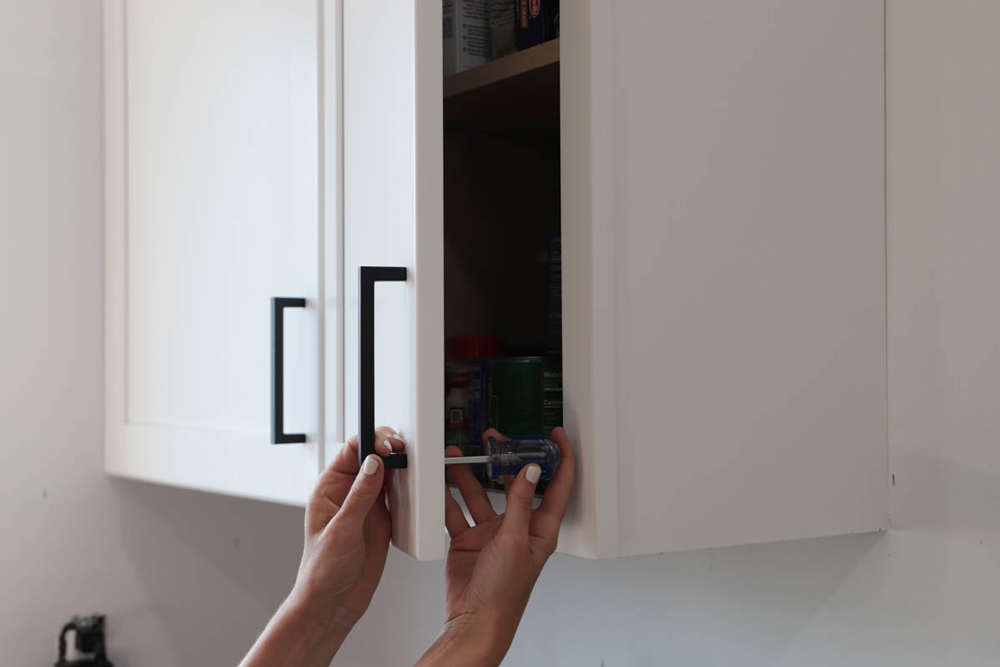 A person installing a black handle on the cabinet door.