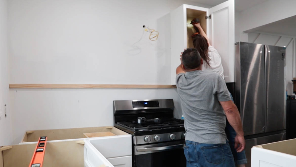 Woman drilling, installing a cabinet into a wall as a man holds the cabinet to support.