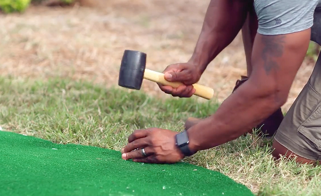 A person using a mallet and pegs to nail down synthetic turf for a putting green.