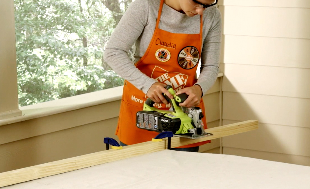 A woman wearing safety goggles and an orange apron uses a circular saw to make cuts in the wood for the DIY blanket ladder.