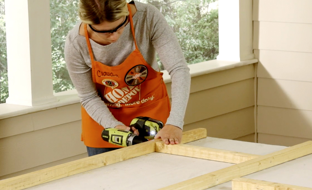 A woman wearing safety goggles and an orange apron uses a drill to attach the first rung of a DIY blanket ladder.