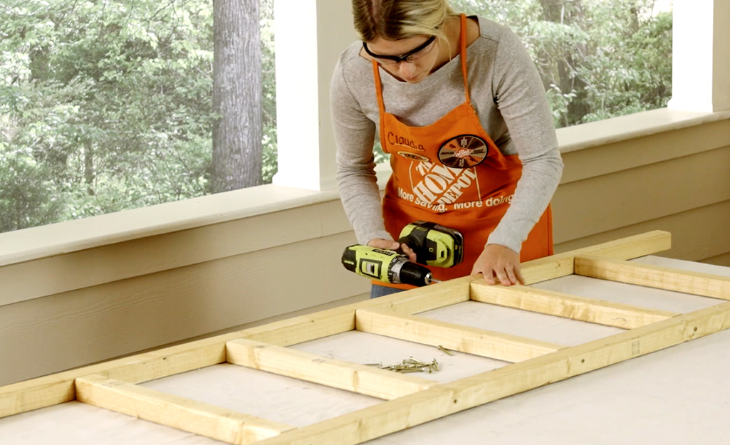 A woman wearing safety goggles and an orange apron uses a drill to attach the rungs of a DIY blanket ladder.