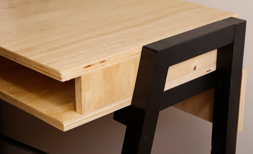 Black legs are attached to the side of a wooden box that serves as the body of a DIY desk. Black legs are attached to the side of a wooden box that serves as the body of a DIY desk.