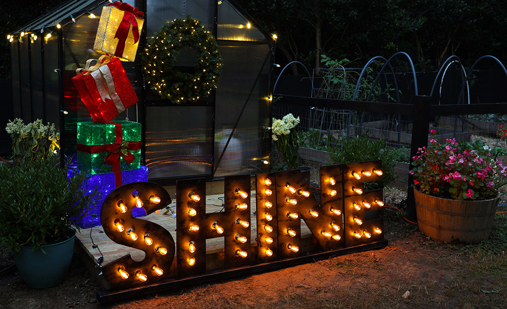 An illuminated faux marquee letter sign reading "SHINE" in front of a greenhouse decorated for Christmas.