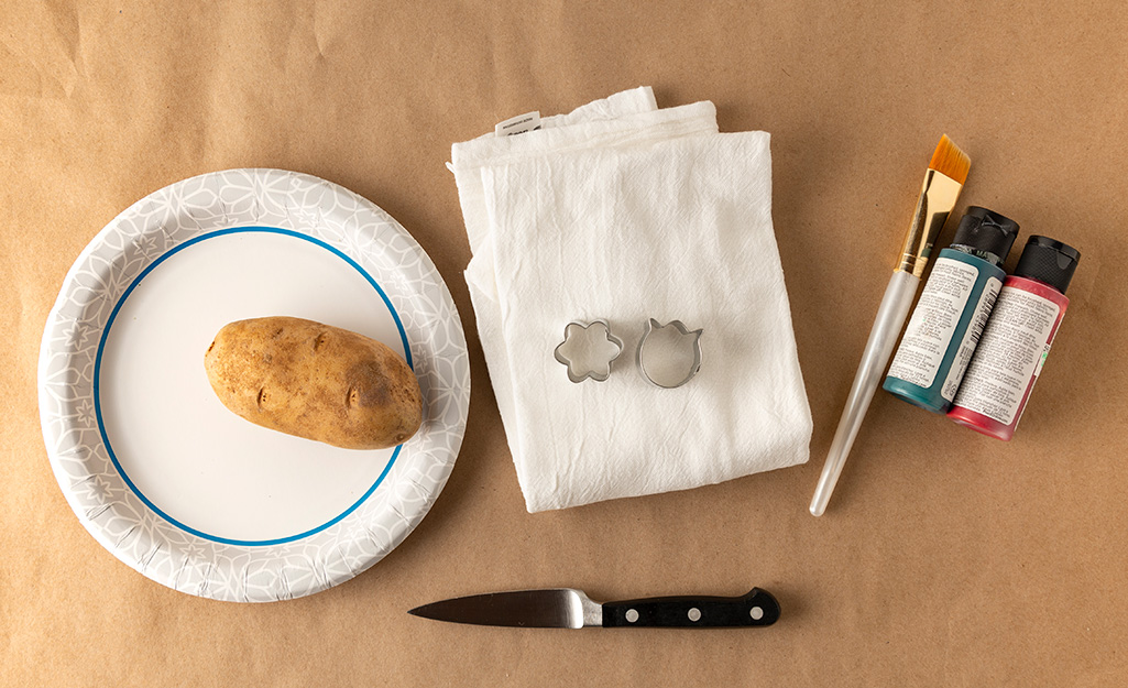 A potato on a paper plate sits next to a knife, a cookie cutter, tea towels, a small paint brush and two bottles of paint.