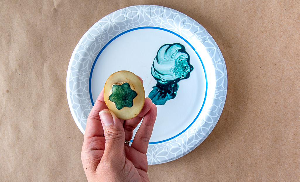 A person holds a potato stamp dipped in paint above a paper plate.