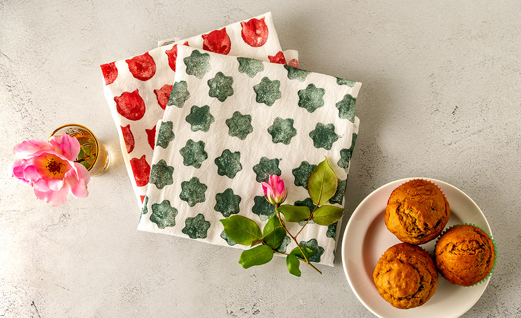 Two folded tea towels stamped with custom patterns lay next to flowers and a plate of muffins.