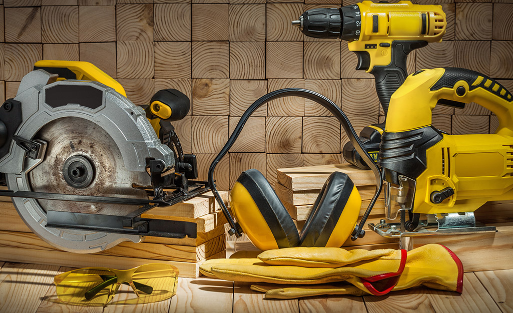 A circular saw, power drill, jigsaw, ear protection and leather work gloves lay on a work surface.