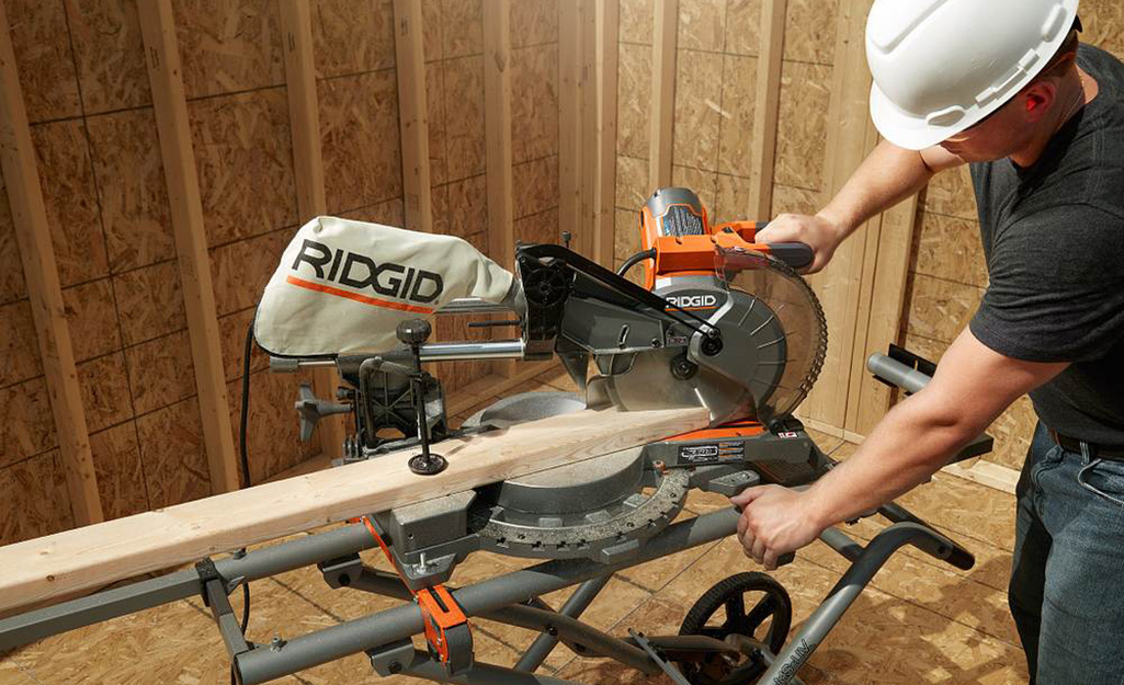 A person wearing a white hard hat uses a miter saw to cut an angle into the end of a board.
