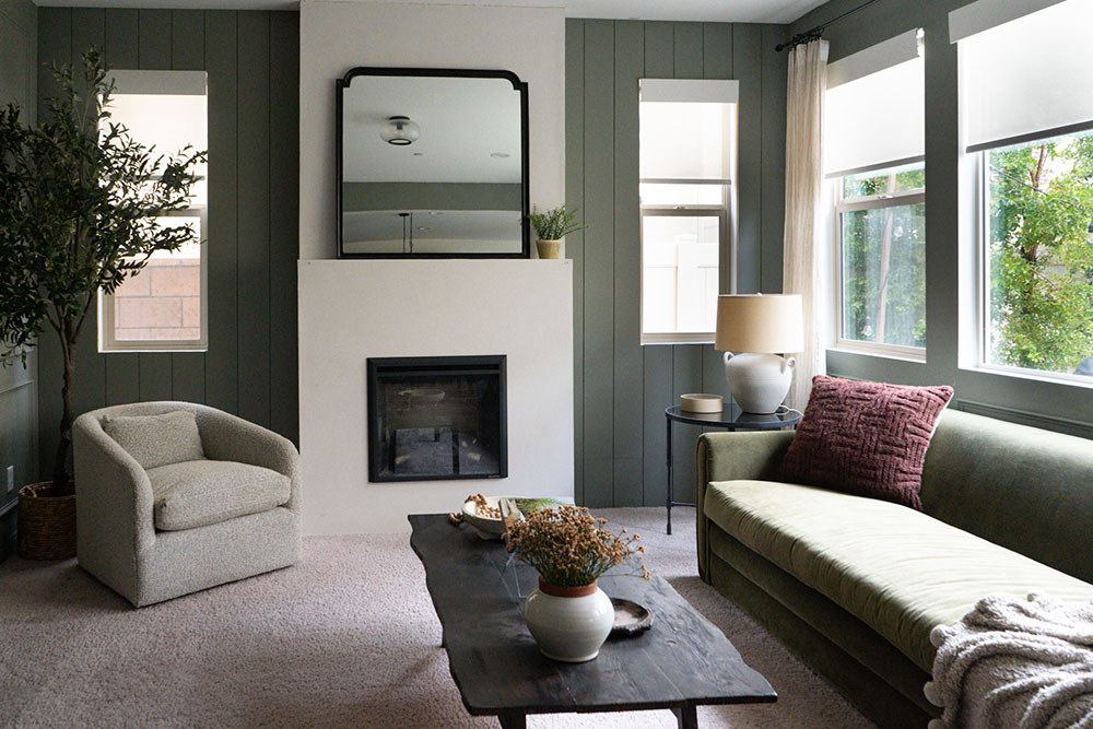 A room with a couch, wood table, fireplace with 4 windows with white window treatments. A room with a couch, wood table, fireplace with 4 windows with white window treatments.