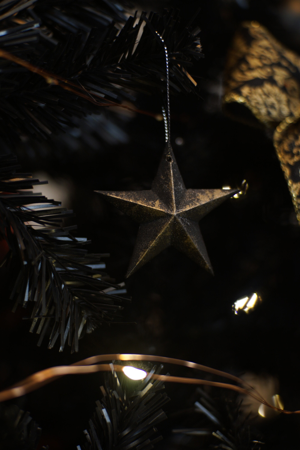 Close-up of a dark star ornament hanging on a black Christmas tree with lights.