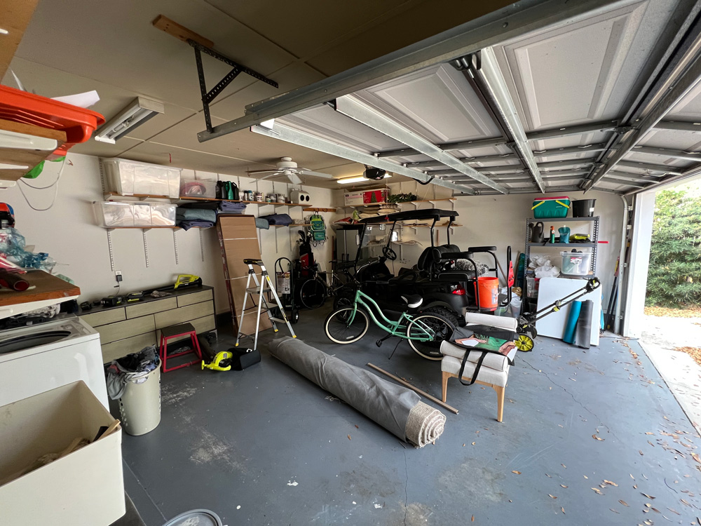 Wide angle photo of inside a cluttered garage with a bike, golf cart, rug, step ladder and more.
