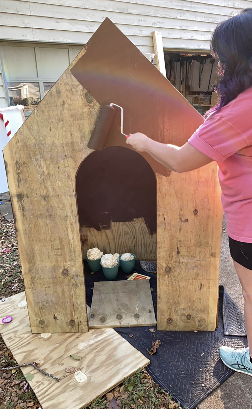 A woman painting the outside of a wooden gingerbread house brown.