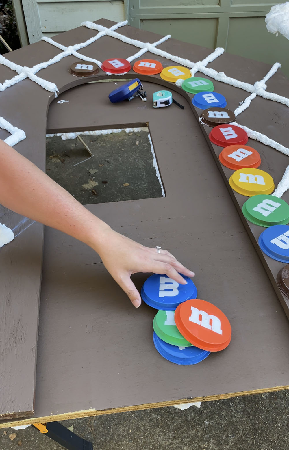 A woman applying fake, custom M&M’s using construction adhesive to a wooden gingerbread house.