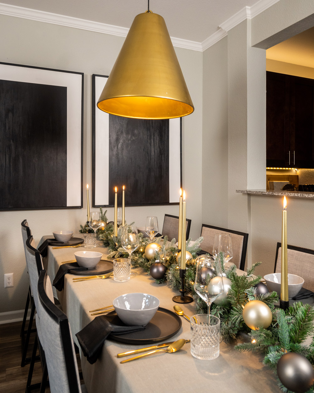 Fully set holiday table with garland centerpiece with gold and grey ornaments, lit candles, and white and grey dinnerware with a light grey tablecloth and gold pendant light above. Fully set holiday table with garland centerpiece with gold and grey ornaments, lit candles, and white and grey dinnerware with a light grey tablecloth and gold pendant light above.