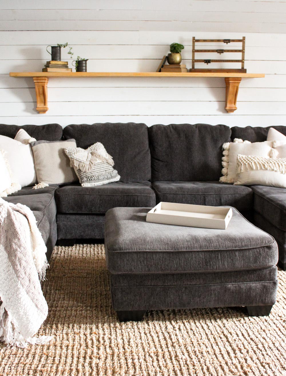 A gray sectional sits in front of a white shiplap wall with a wood shelf.