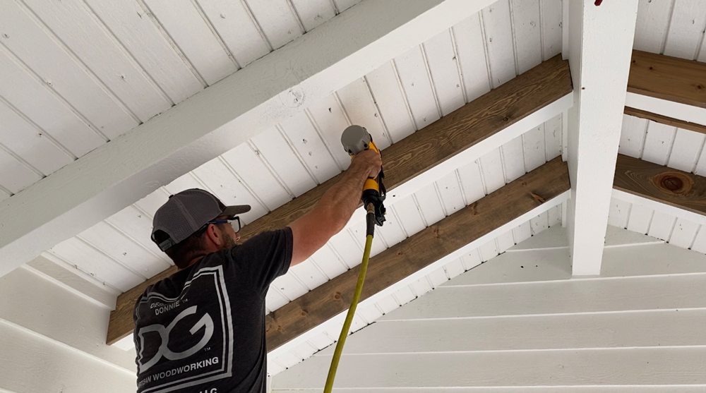 A man using a nailer to nail down the ceiling beams.